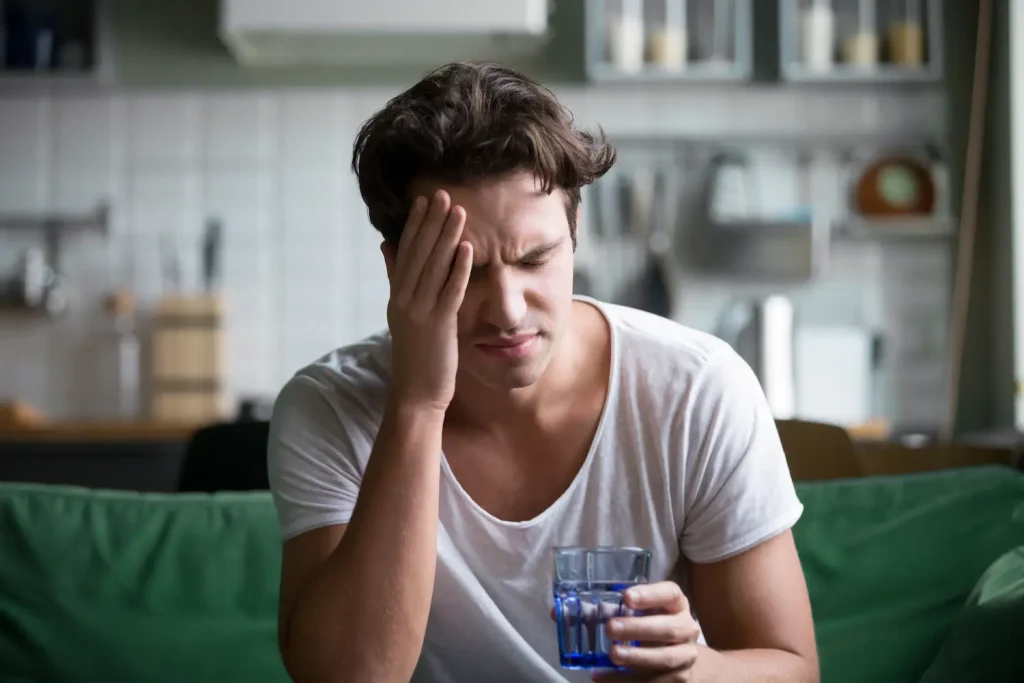 A man sitting on a couch, holding his head in pain, indicating he is suffering from a migraine.